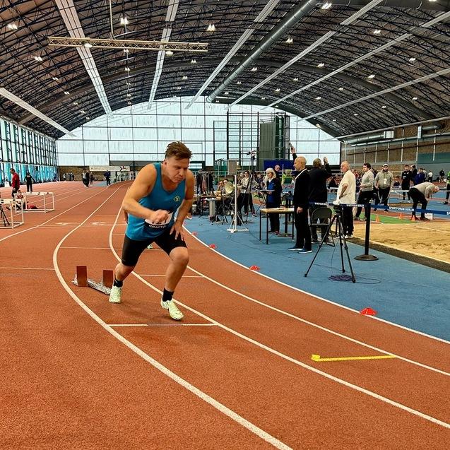 Ein Athlet startet sprintend auf einer Leichtathletikbahn in einer Sporthalle. Zuschauer und Geräte im Hintergrund.