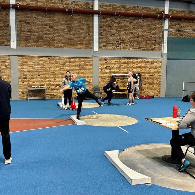 Athlet wirft in blauer Sportkleidung eine Kugel in einer Sporthalle, während Zuschauer und Kampfrichter beobachten.