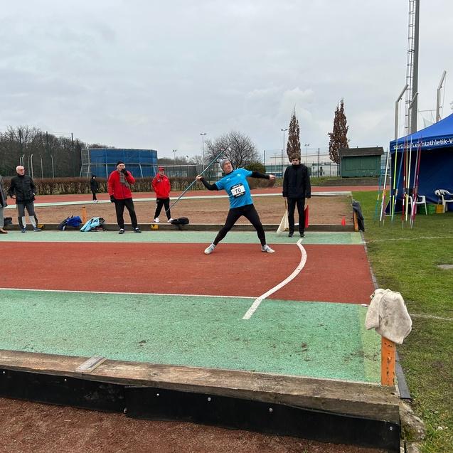 Athlet wirft eine Speer auf einem Wettkampfsportplatz, während Zuschauer und Trainer in der Nähe stehen.