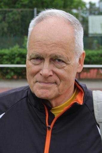 Älterer Mann mit kurzen grauen Haaren trägt eine Sportjacke und blickt direkt in die Kamera, Sportanlage im Hintergrund.