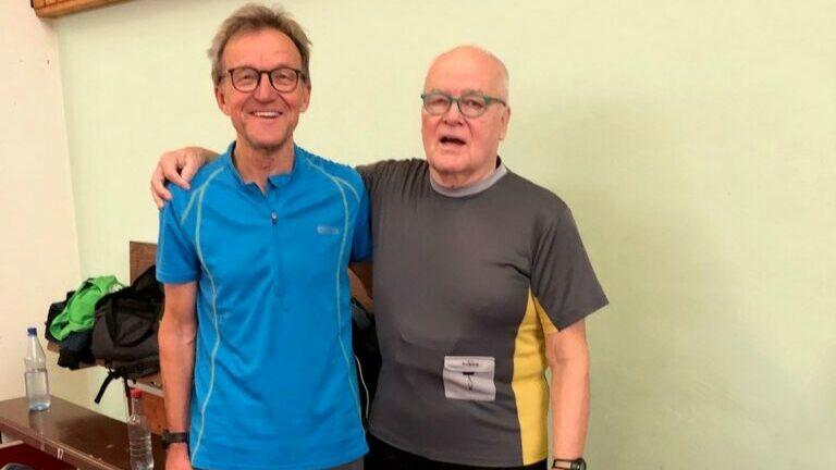 Zwei ältere Männer in Sportkleidung lächeln nebeneinander in einer Turnhalle, im Hintergrund Sportutensilien.