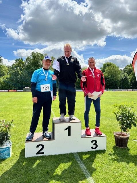 Drei Medaillengewinner stehen auf dem Siegertreppchen im Freien; der Erste trägt eine Medaille und hält ein Zertifikat.