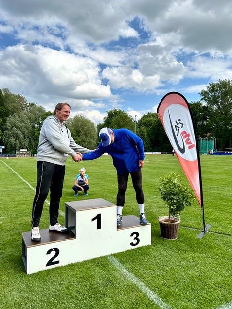 Zwei Männer auf einem Siegertreppchen in einem Stadion, während ein Kind im Hintergrund sitzt und zuschaut.