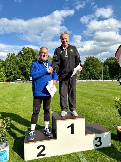 Zwei Männer stehen auf dem Siegerpodest, der erste winkt, der zweite reicht ihm die Hand und hält Urkunden.