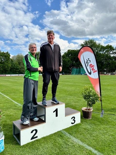 Zwei Männer stehen auf einem Siegerpodest im Freien, umgeben von grünem Gras und einem Wettkampf-Flaggen.