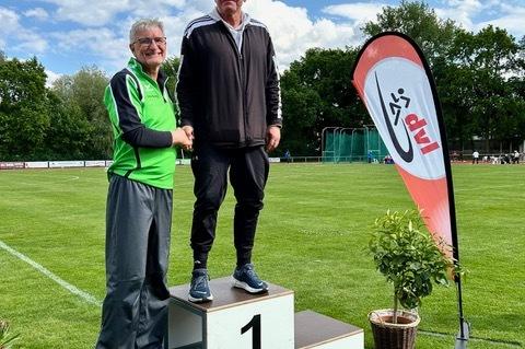 Zwei M&auml;nner stehen auf einem Siegerpodest im Freien, umgeben von gr&uuml;nem Gras und einem Wettkampf-Flaggen.