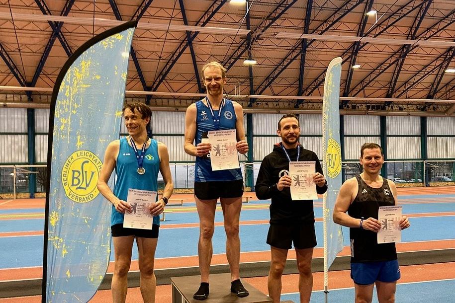 Vier m&auml;nnliche Athleten auf dem Siegertreppchen mit Urkunden in der Hand, im Hintergrund eine Sporthalle und ein Vereinsbanner.