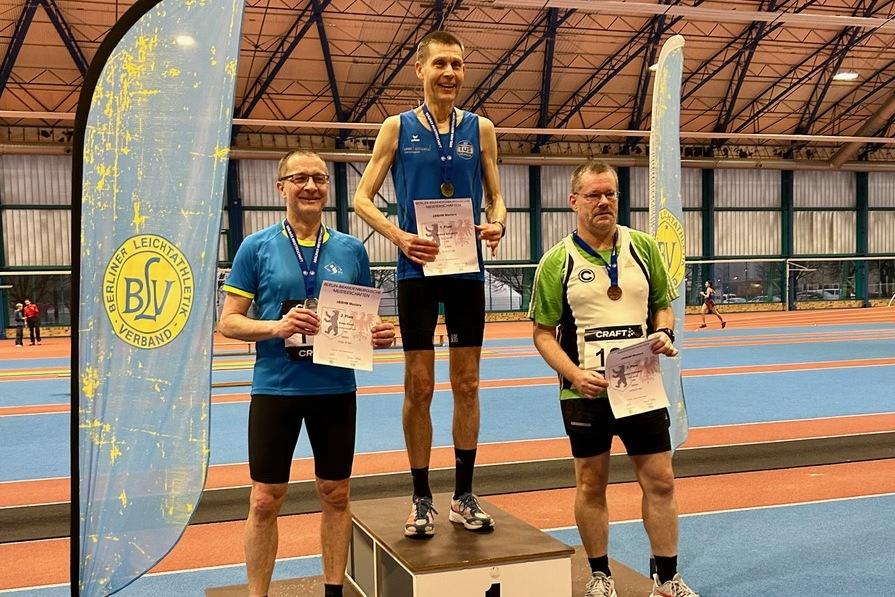 Drei hergestellte Sportler auf dem Siegertreppchen halten Urkunden; ein Banner des Berliner Leichtathletikverbands im Hintergrund.