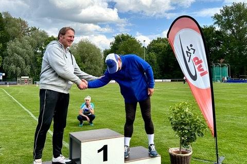 Zwei M&auml;nner auf einem Siegertreppchen in einem Stadion, w&auml;hrend ein Kind im Hintergrund sitzt und zuschaut.