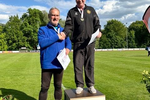 Zwei M&auml;nner stehen auf dem Siegerpodest, der erste winkt, der zweite reicht ihm die Hand und h&auml;lt Urkunden.