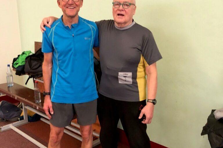 Zwei &auml;ltere M&auml;nner in Sportkleidung l&auml;cheln nebeneinander in einer Turnhalle, im Hintergrund Sportutensilien.