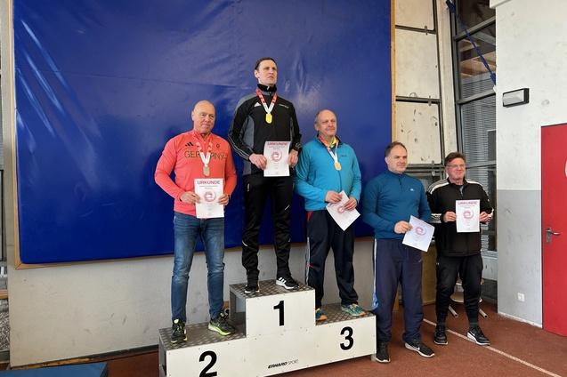 Fünf Sportler stehen auf dem Siegerpodest, halten Urkunden und Medaillen in der Hand. Blaues Paspellierungs-Hintergrund.