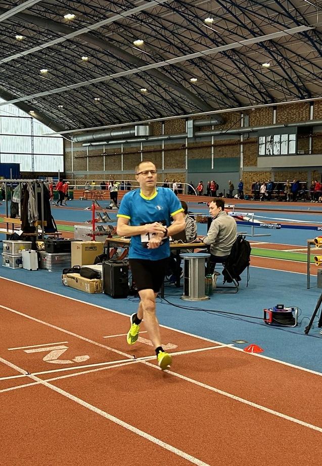 Ein Athlet in blauer Sportkleidung läuft auf einer Laufbahn in einer Sporthalle während eines Wettkampfs.