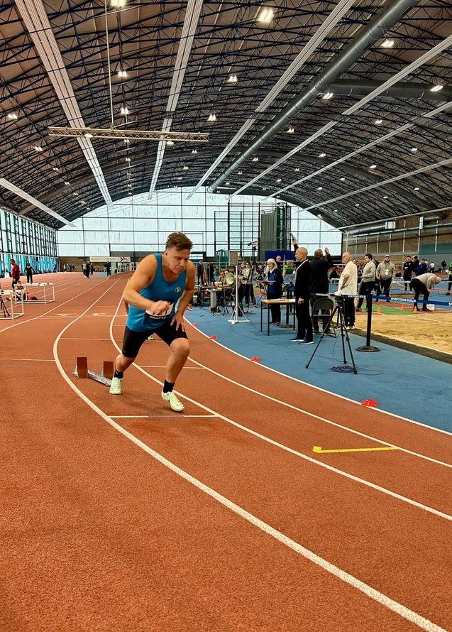 Ein Athlet startet sprintend auf einer Leichtathletikbahn in einer Sporthalle. Zuschauer und Geräte im Hintergrund.