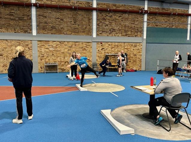 Athlet wirft in blauer Sportkleidung eine Kugel in einer Sporthalle, während Zuschauer und Kampfrichter beobachten.