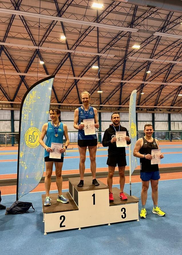 Vier männliche Athleten auf dem Siegertreppchen mit Urkunden in der Hand, im Hintergrund eine Sporthalle und ein Vereinsbanner.