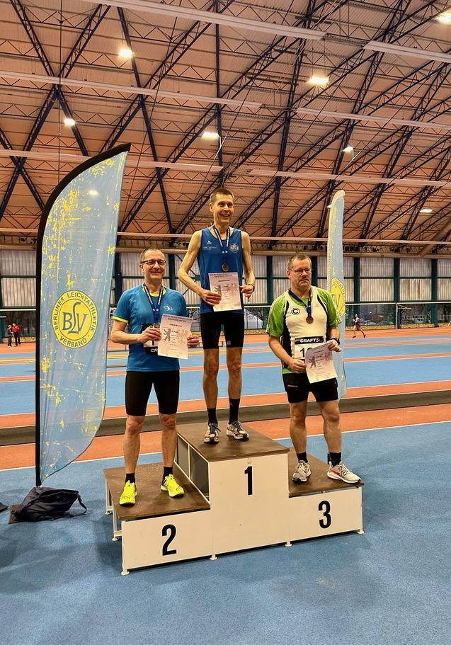 Drei hergestellte Sportler auf dem Siegertreppchen halten Urkunden; ein Banner des Berliner Leichtathletikverbands im Hintergrund.