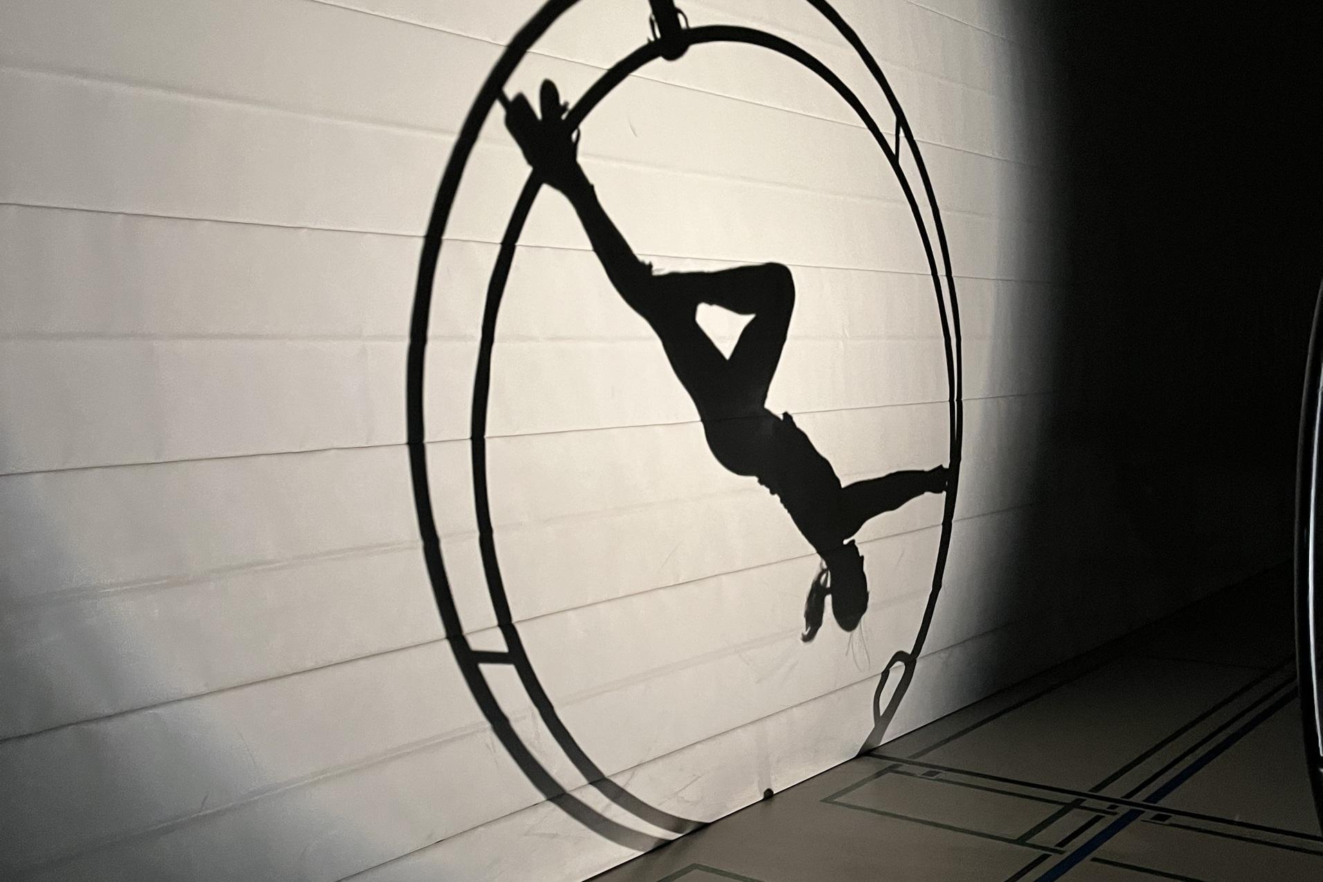 Schatten einer Person, die in einem Zirkusrad auf einer Wand schwebt, beleuchtet von sanftem Licht.