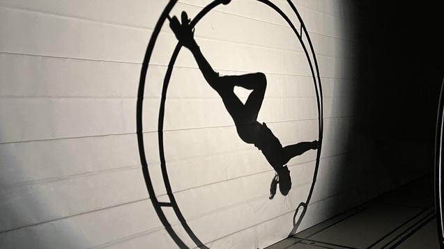 Schatten einer Person, die in einem Zirkusrad auf einer Wand schwebt, beleuchtet von sanftem Licht.