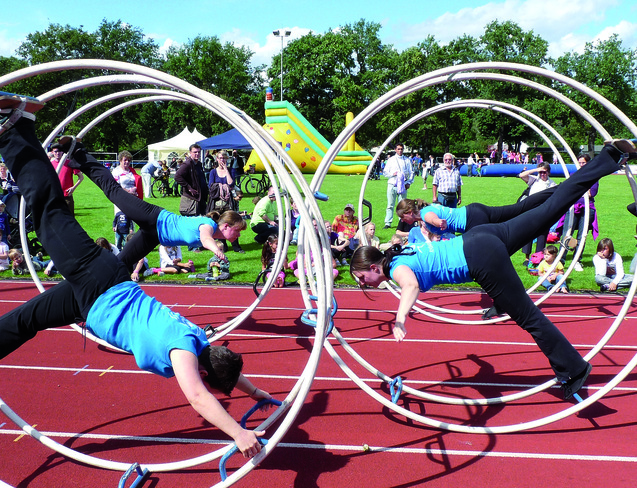 Drei Sportler in blauen T-Shirts absolvieren akrobatische &Uuml;bungen in gro&szlig;en Holzreifen auf einer Laufbahn.