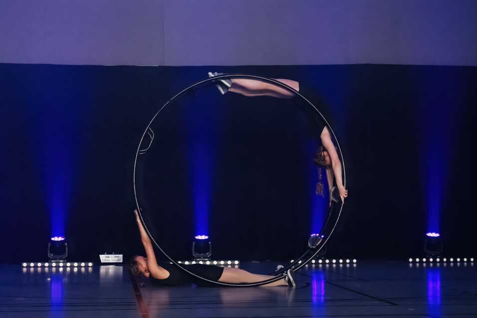 Zwei Artistinnen bringen eine synchronisierte Darbietung mit einem großem Ring auf einer Bühne mit blauer Beleuchtung.
