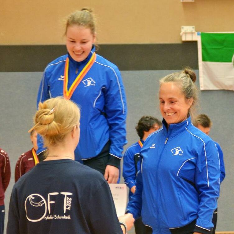 Zwei Sportlerinnen in blauen Trainingsjacken auf dem Siegertreppchen, eine erhält eine Urkunde von einer anderen.