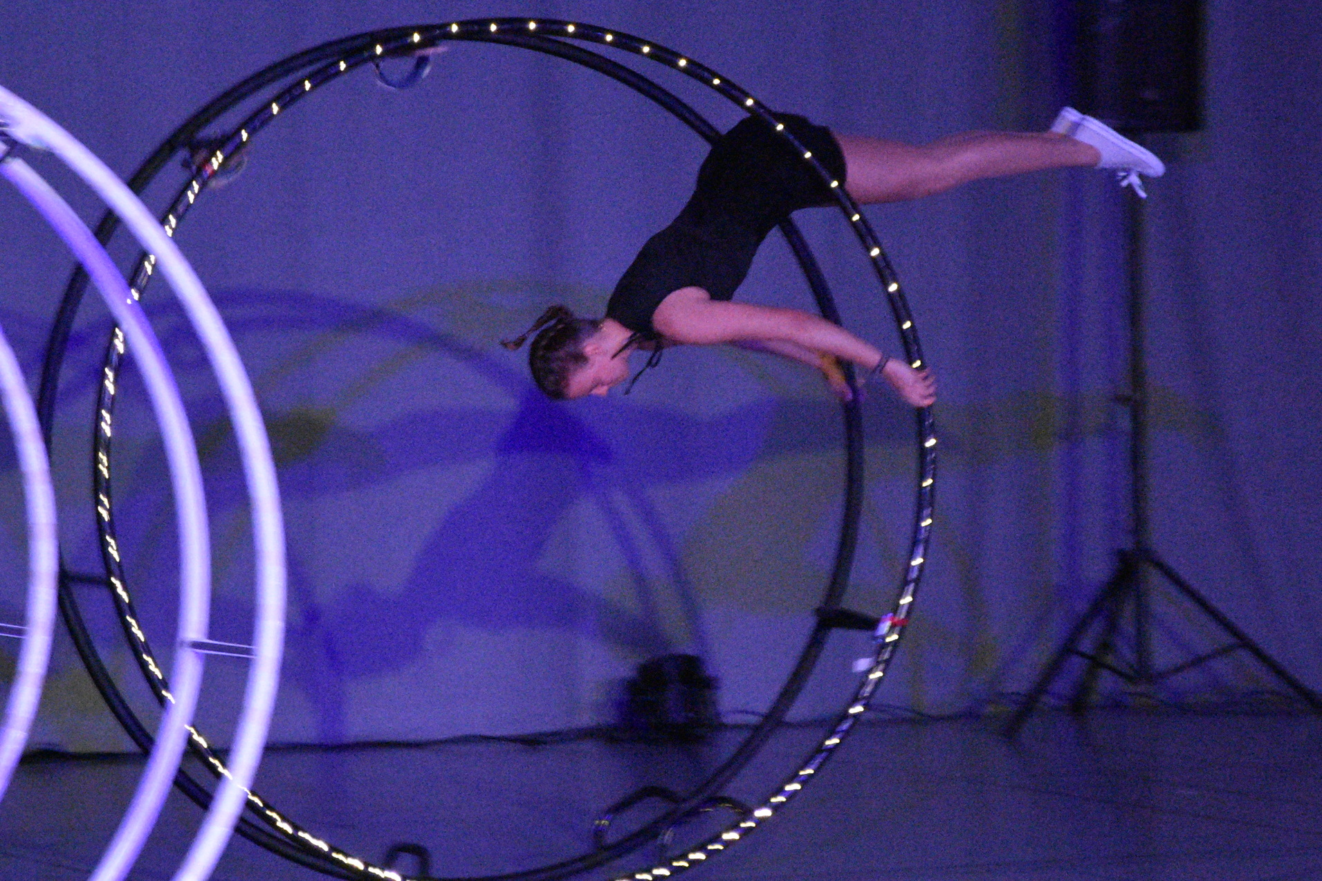 Artistikende Frau schwingt an einem gro&szlig;en Hula-Hoop-Reifen auf einer B&uuml;hne mit Lichtern und dunklem Hintergrund.