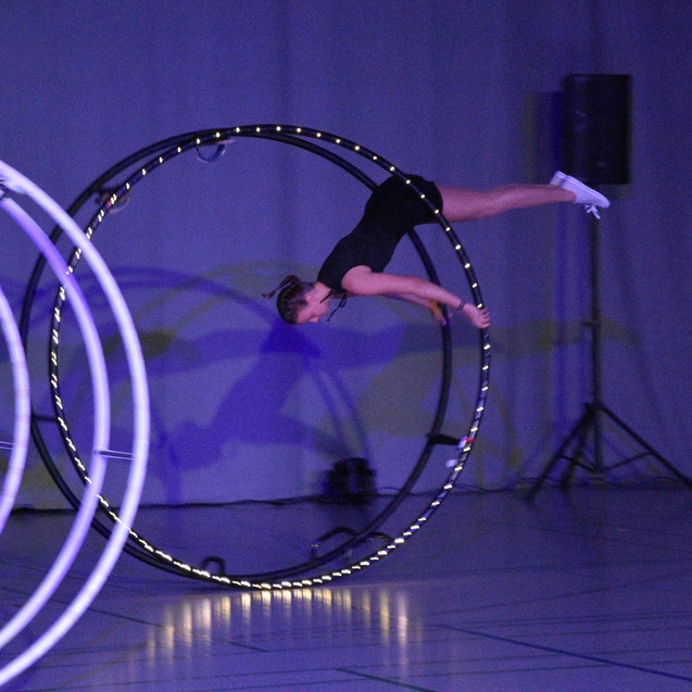 Artistikende Frau schwingt an einem gro&szlig;en Hula-Hoop-Reifen auf einer B&uuml;hne mit Lichtern und dunklem Hintergrund.