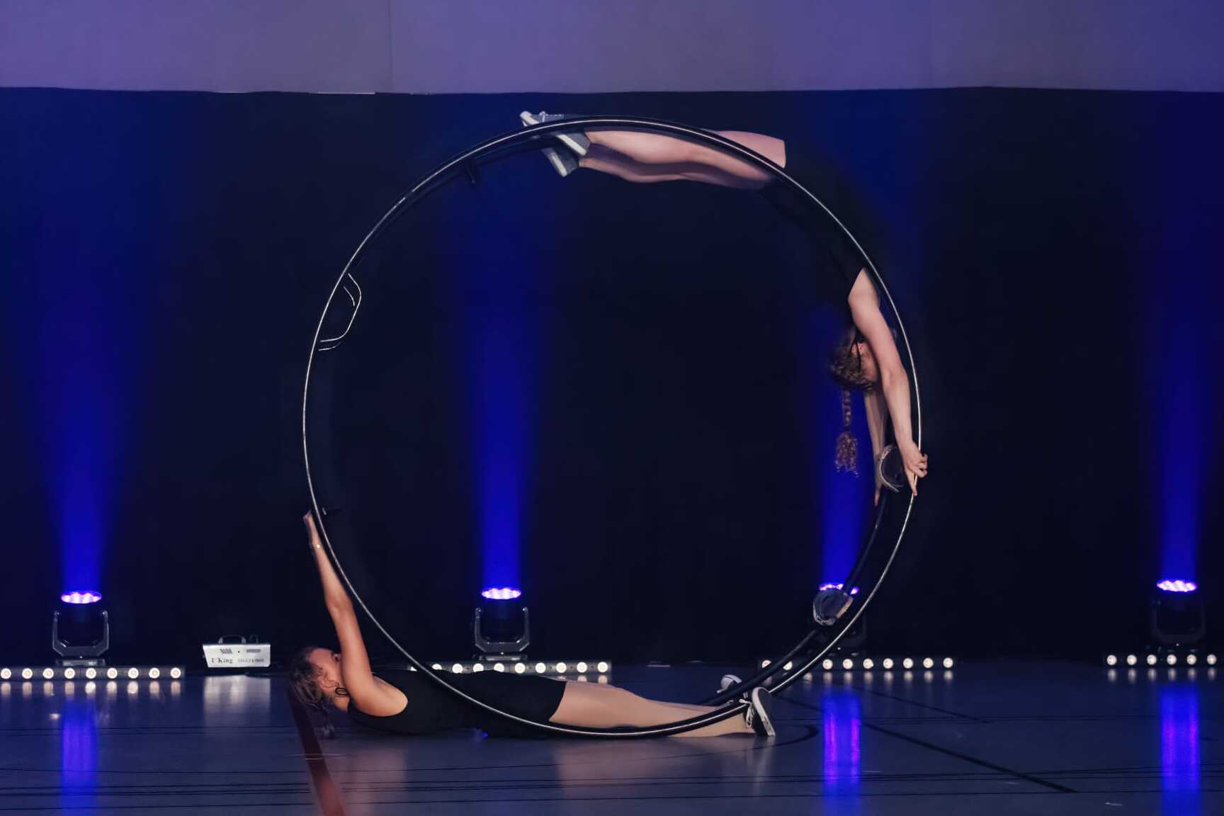 Zwei Artistinnen bringen eine synchronisierte Darbietung mit einem großem Ring auf einer Bühne mit blauer Beleuchtung.