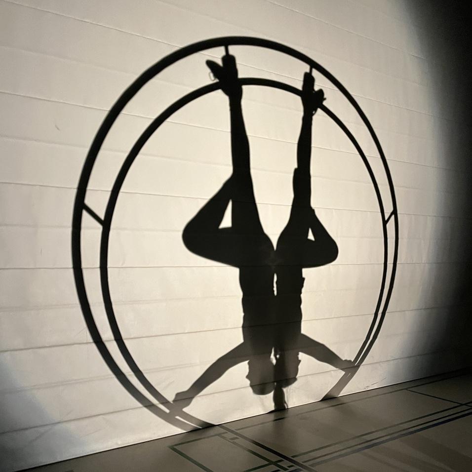 Schatten einer Person, die kopf&uuml;ber in einem gro&szlig;en Kreis h&auml;ngt, vor einer hellen Wand in einer Sporthalle.