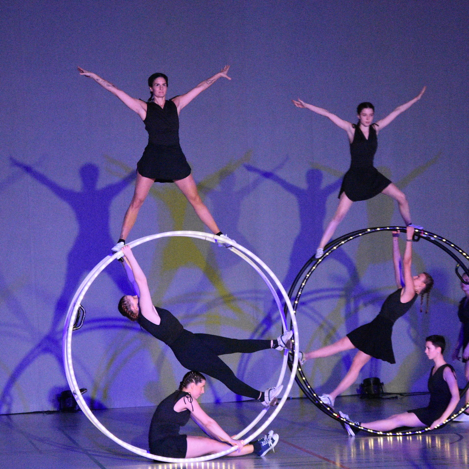 Vier Artistinnen in Schwarz balancieren in verschiedenen Positionen auf drei großen Reifen auf einer Bühnenfläche.