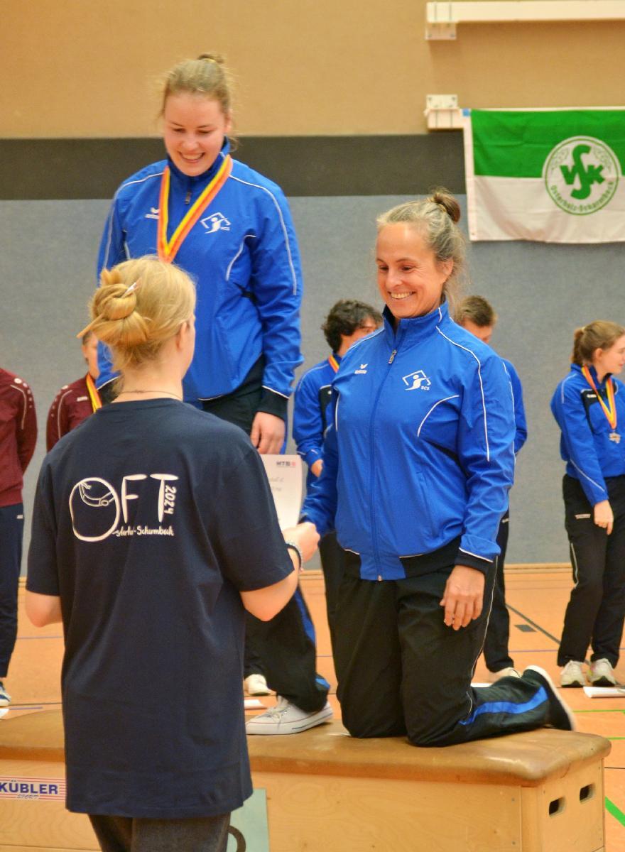 Zwei Sportlerinnen in blauen Trainingsjacken auf dem Siegertreppchen, eine erhält eine Urkunde von einer anderen.