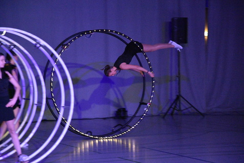 Artistikende Frau schwingt an einem großen Hula-Hoop-Reifen auf einer Bühne mit Lichtern und dunklem Hintergrund.