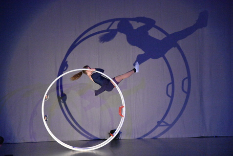 Akrobatin schwingt in einem Ring, während ihr Schatten an der Wand eine dynamische Pose zeigt.