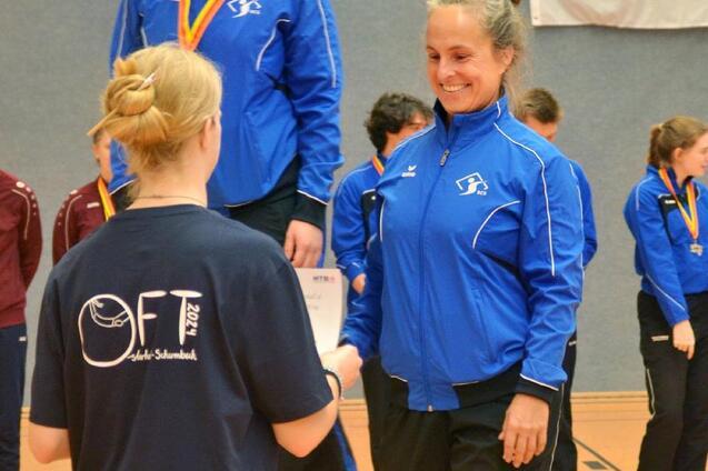Zwei Sportlerinnen in blauen Trainingsjacken auf dem Siegertreppchen, eine erh&auml;lt eine Urkunde von einer anderen.