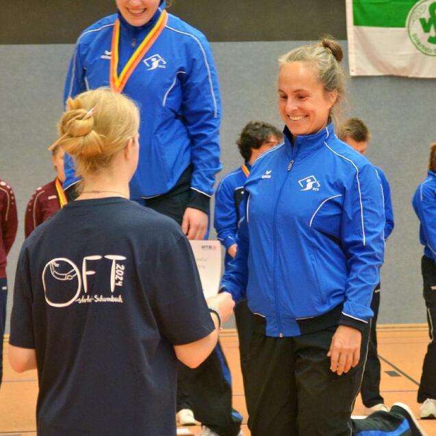 Zwei Sportlerinnen in blauen Trainingsjacken auf dem Siegertreppchen, eine erh&auml;lt eine Urkunde von einer anderen.