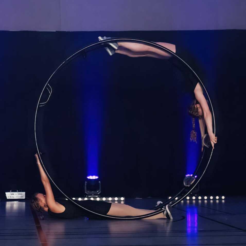 Zwei Artistinnen bringen eine synchronisierte Darbietung mit einem großem Ring auf einer Bühne mit blauer Beleuchtung.