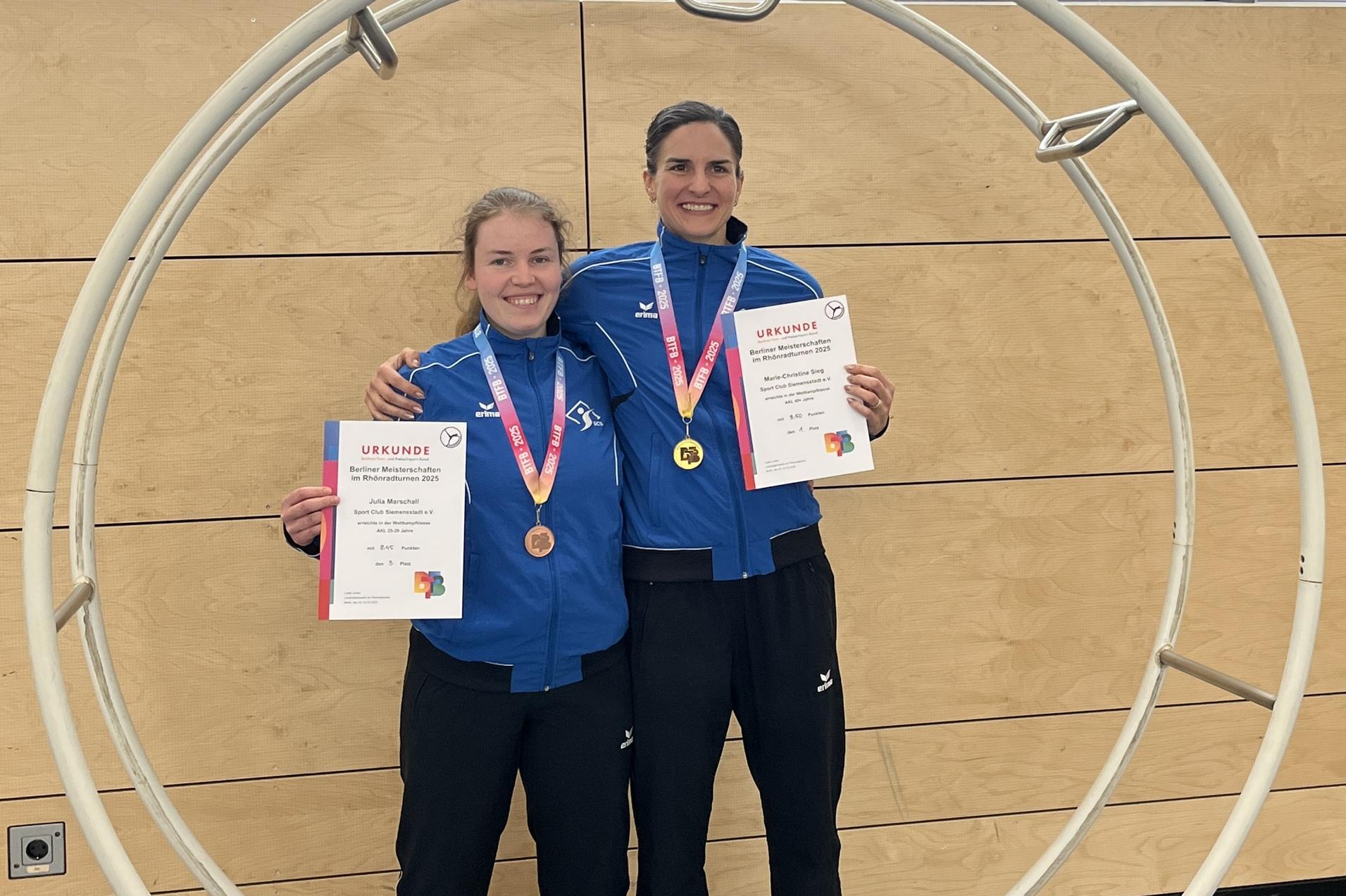Zwei Sportlerinnen in blauen Trainingsanz&uuml;gen stehen l&auml;chelnd in einem gro&szlig;en Ring und halten Urkunden und Medaillen.