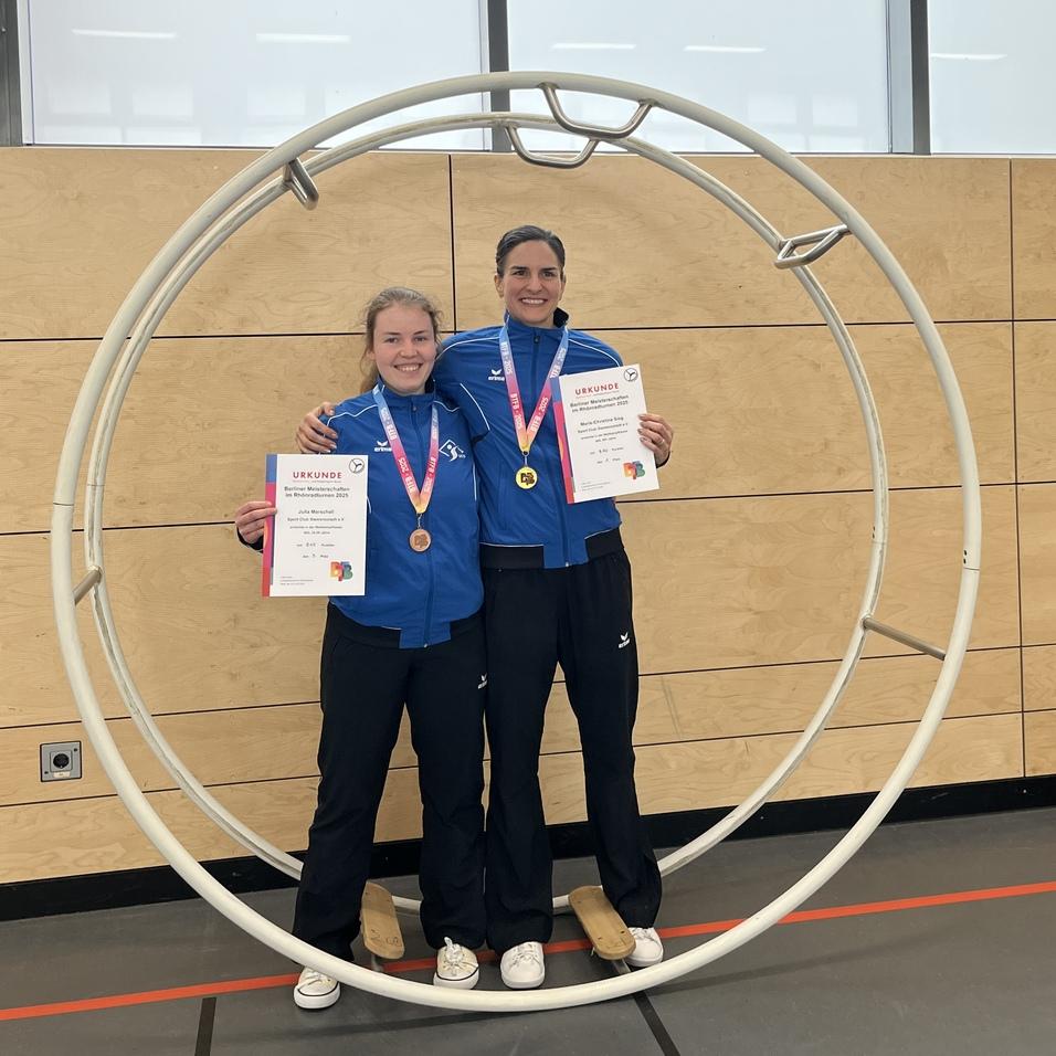 Zwei Sportlerinnen in blauen Trainingsanzügen stehen lächelnd in einem großen Ring und halten Urkunden und Medaillen.