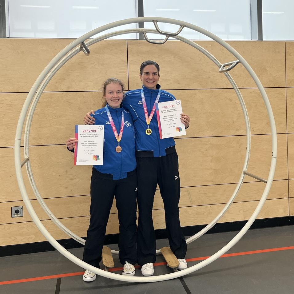 Zwei Sportlerinnen in blauen Trainingsanzügen stehen lächelnd in einem großen Ring und halten Urkunden und Medaillen.