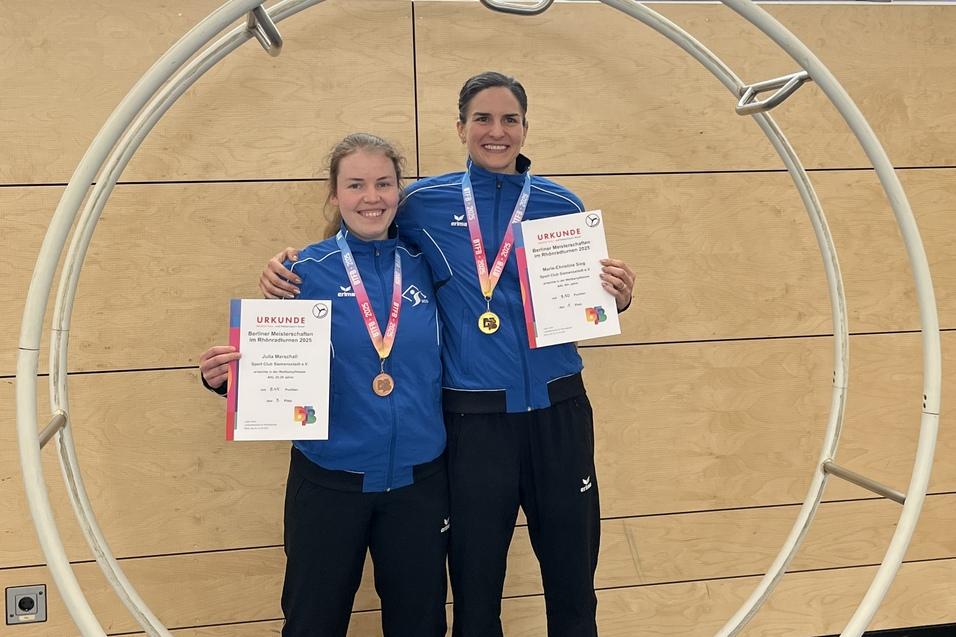 Zwei Sportlerinnen in blauen Trainingsanz&uuml;gen stehen l&auml;chelnd in einem gro&szlig;en Ring und halten Urkunden und Medaillen.