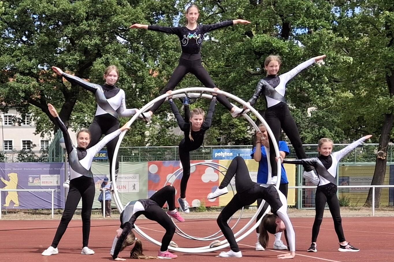 Gruppe von Mädchen in Sportanzügen bildet kunstvolle Pyramide auf einem Sportplatz mit einem großen Reifen im Hintergrund.