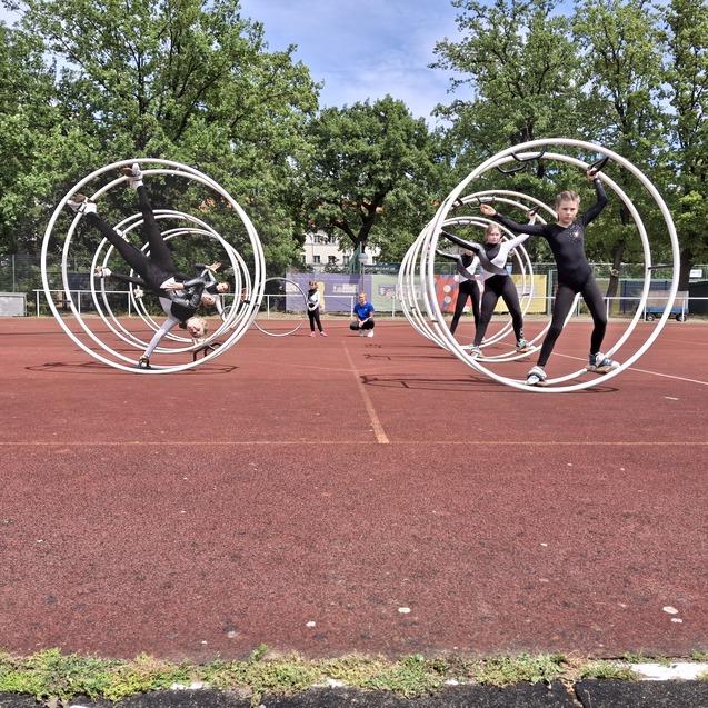 Kinder in Schwarz führen Tricks in großen, weißen Zirkusschwingen auf einem roten Sportplatz aus.