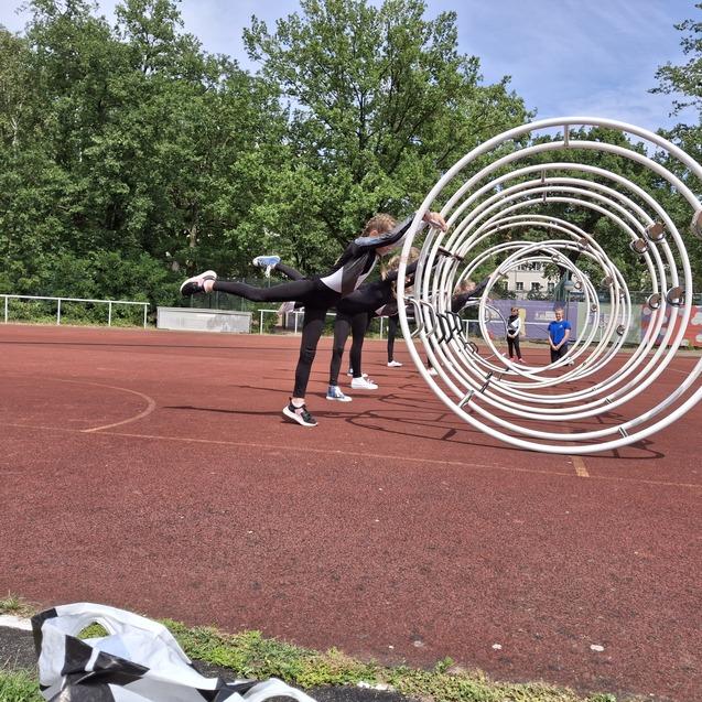 Sportler üben im Freien mit Trainingsgeräten auf einem roten Sportplatz, umgeben von grünen Bäumen und anderen Teilnehmern.