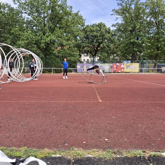 Gruppe von Sportlern trainiert auf einem roten Platz mit großen Reifen; eine Person rollt gerade durch einen Reifen.