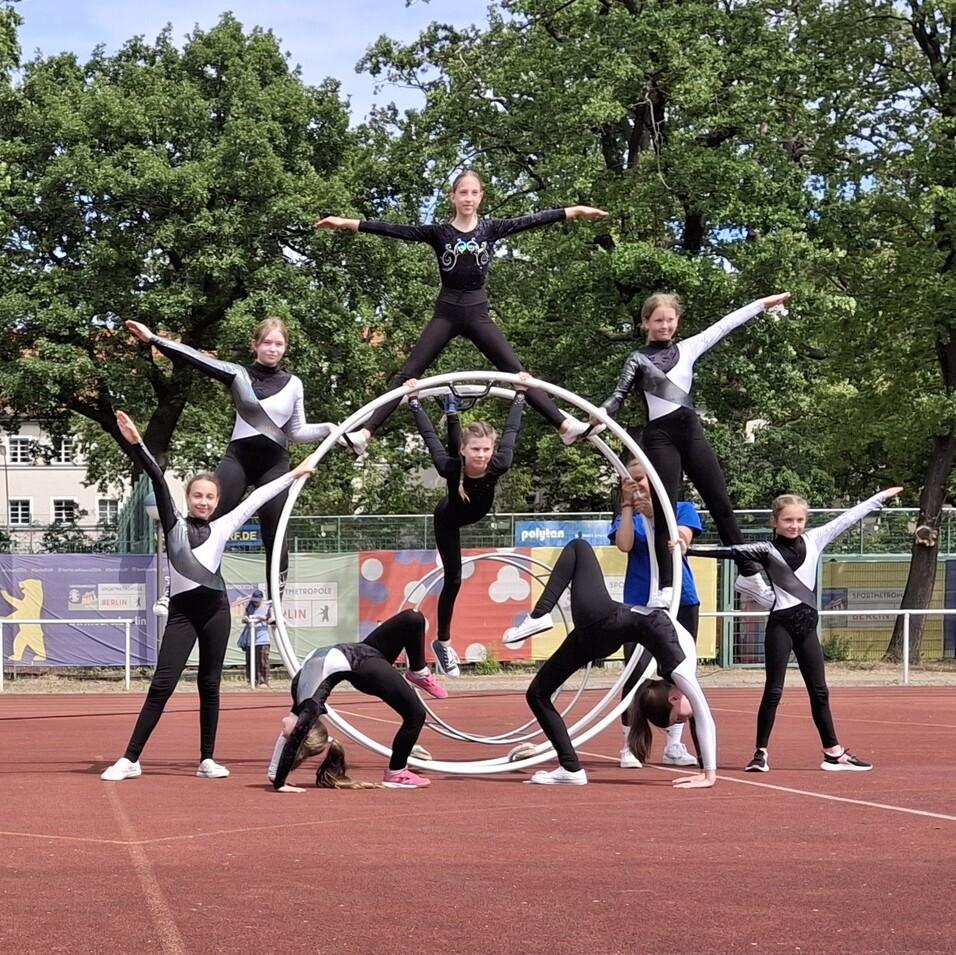 Gruppe von Mädchen in Sportanzügen bildet kunstvolle Pyramide auf einem Sportplatz mit einem großen Reifen im Hintergrund.