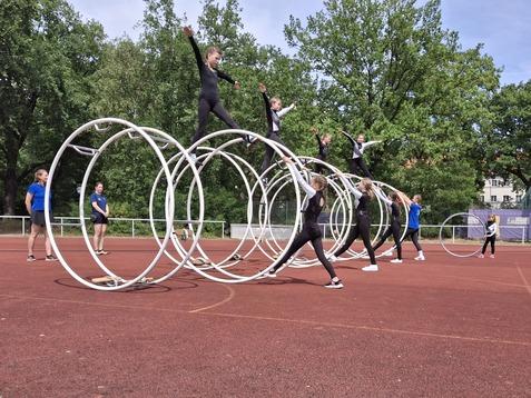 Gruppe von Sportlerinnen, die auf Hoops auf einem roten Sportplatz trainieren, unter Bäumen und blauem Himmel.