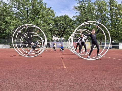 Kinder in Schwarz führen Tricks in großen, weißen Zirkusschwingen auf einem roten Sportplatz aus.