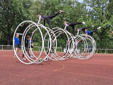 Gruppen von Kindern turnen kreativ mit großen Reifen auf einem sportlichen Platz mit rotem Belag.
