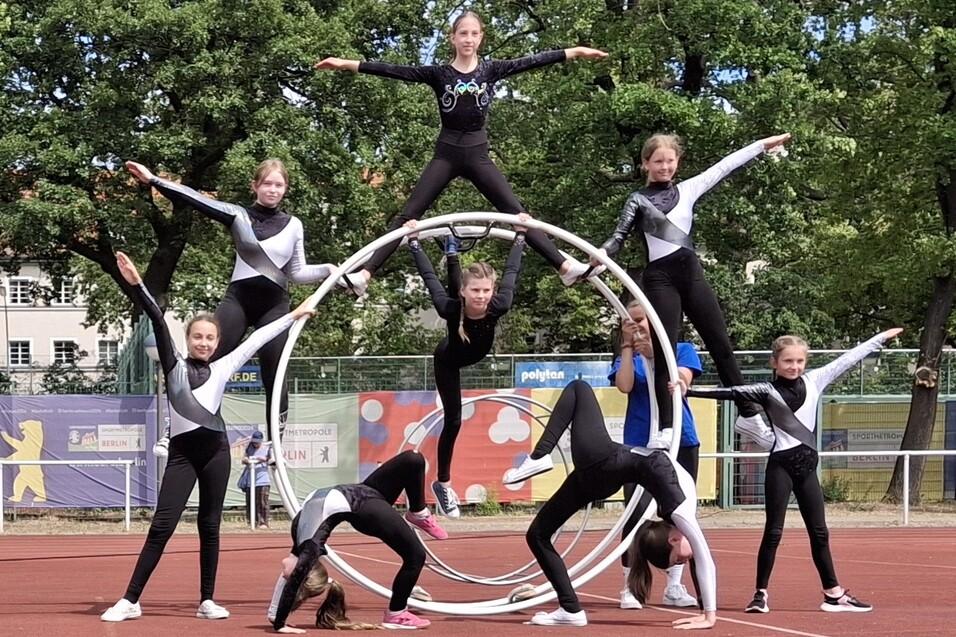 Gruppe von M&auml;dchen in Sportanz&uuml;gen bildet kunstvolle Pyramide auf einem Sportplatz mit einem gro&szlig;en Reifen im Hintergrund.
